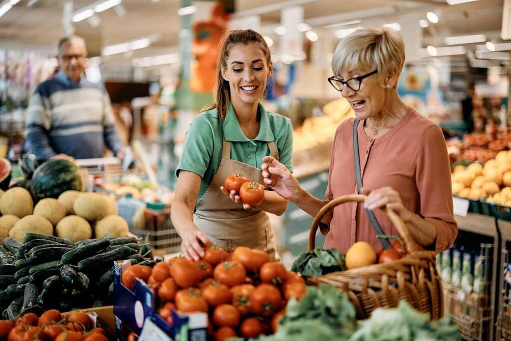 funcionária ajudando senhora a escolher itens no supermercado, representando as compras feitas com os valores da tabela do salário mínimo