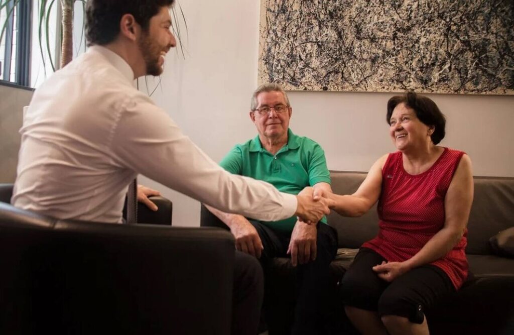 Atendimento jurídico a casal de idosos sobre Isenção de Imposto de Renda por câncer curado, com advogado explicando direitos e orientações.