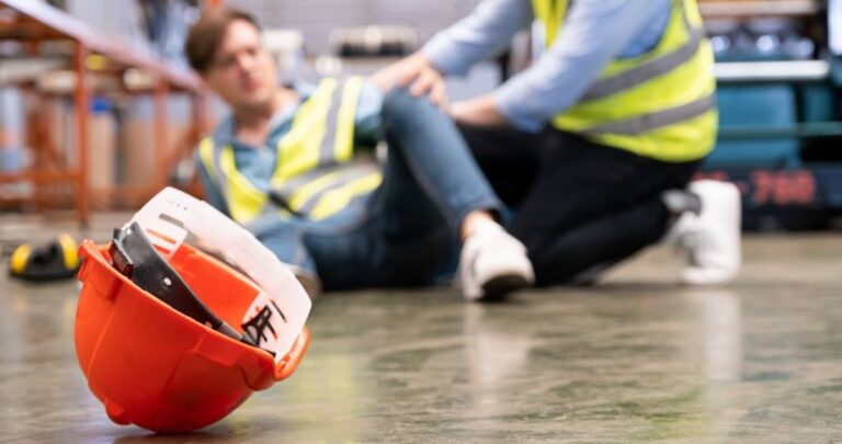Capacete de proteção laranja caído no chão de uma fábrica em primeiro plano, com um trabalhador acidentado ao fundo recebendo auxílio médico de forma desfocada.