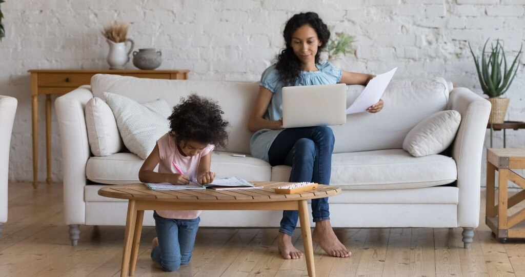 Mãe brasileira trabalhando em um notebook no sofá de casa ao lado da filha, pesquisando sobre o salário maternidade aprovado, como receber os valores previdenciários e conferindo documentos.